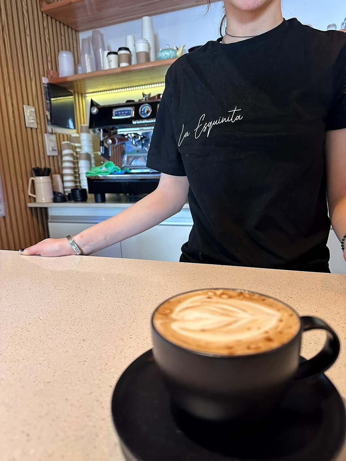 Barista de La Esquinita con remera de la casa y un flat white con latte art